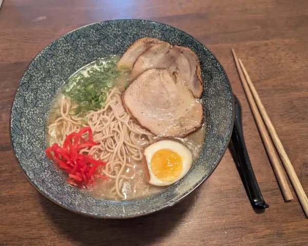 Miseczka z ramen: makaron, plastry mięsa, jajko na pół, zielona cebulka i czerwone przyprawy do ramen. Obok pałeczki.