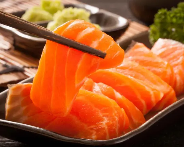 Sashimi co to? Świeże, pomarańczowe kawałki łososia podniesione pałeczkami, obok zielonej pasty wasabi i natki pietruszki.