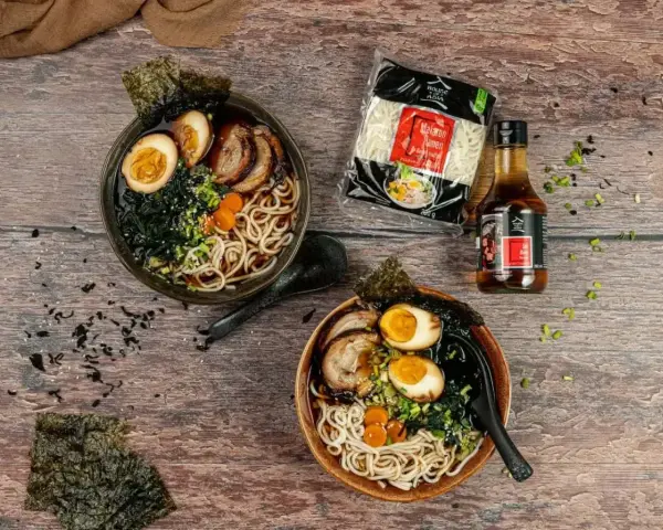 Dwie miski aromatycznego makaronu ramen z sosem sojowym, jajkiem, mięsem i wodorostami. Obok opakowanie makaronu i butelka sosu.