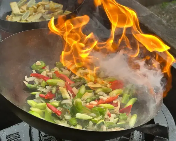 Warzywa w woku płoną w ogniu, tworząc aromatyczny stir fry. W tle widać drugie danie.