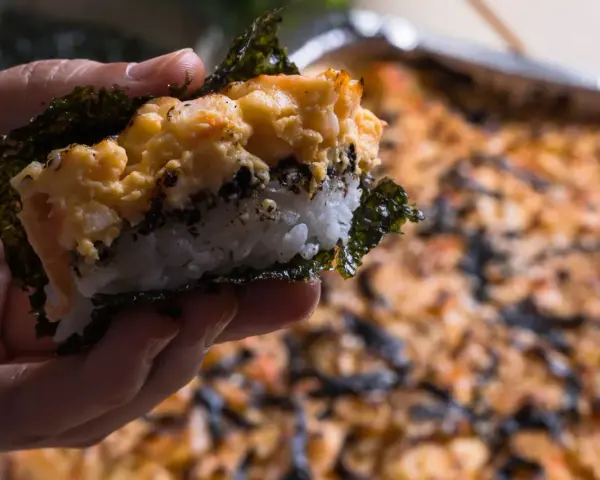 Dłoń trzyma kawałek sushi bake z ryżem, nori i zapiekanym farszem.