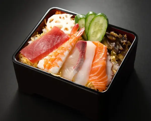 Chirashi zushi: kolorowe plastry ryb (tuńczyk, łosoś, biała ryba), krewetka, plasterki ogórka i lotosu na ryżu.