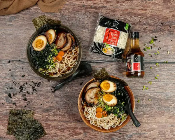 Dwa miski aromatycznego ramenu z boczkiem, jajkiem i nori. Obok opakowanie makaronu i sosu.