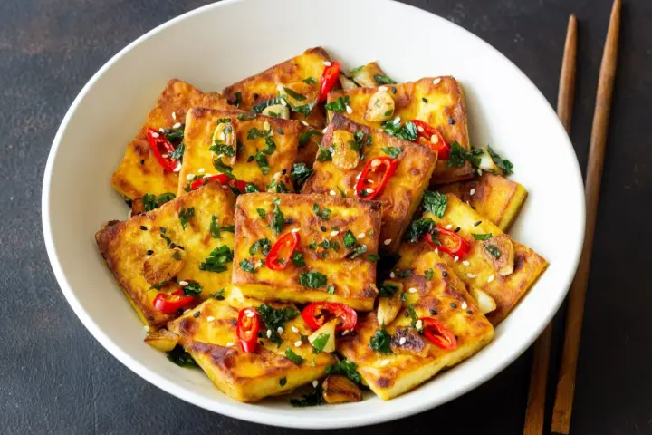 Chrupiące tofu w tempurze, posypane sezamem, chili i natką pietruszki, podane w białej misce z pałeczkami.