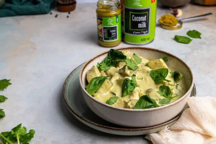 Aksamitny kurczak w zielonym curry, udekorowany świeżym szpinakiem i kolendrą. Obok puszka mleka kokosowego i słoiczek pasty curry.