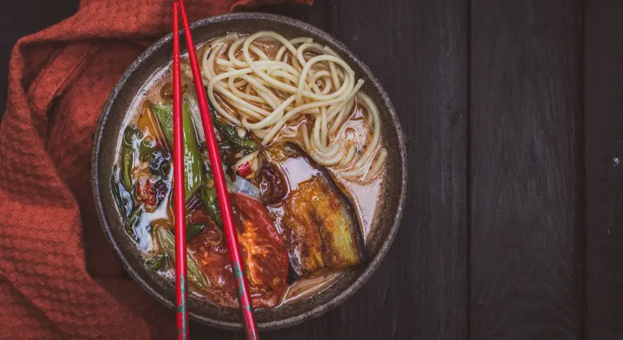 A miseczce z aromatycznym ramen wegetariańskim, pełnej makaronu, grillowanego bakłażana i pomidorów, spoczywają czerwone pałeczki.