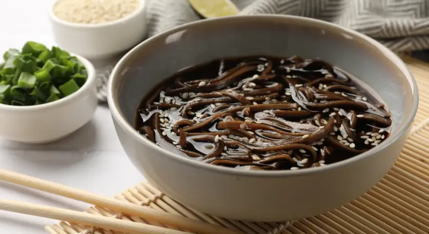 Miseczka z makaronem soba w sosie, posypana sezamem. Obok miseczki z posiekanym szczypiorkiem i sezamem. Zdrowy posiłek, mało makaron soba kcal.