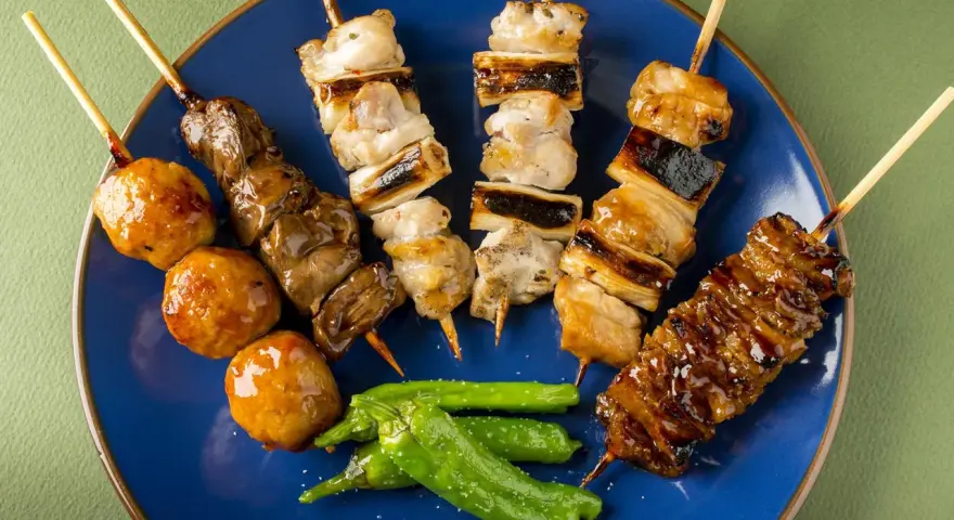 Różnorodne szaszłyki kurczak yakitori: mięso, wątróbka, kulki mięsne i warzywa na niebieskim talerzu.