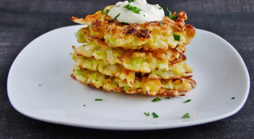 Stos placków okonomiyaki, polanych śmietaną i posypanych natką pietruszki, na białym talerzu.