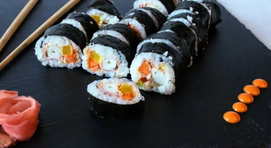 Roladki sushi z surimi, ryżem i warzywami na czarnej desce, obok pałeczki i różowy imbir.