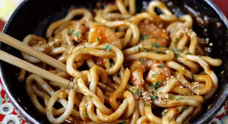 Pyszne udon z krewetkami w sosie, posypane sezamem i natką pietruszki. Danie w czarnej misce, gotowe do jedzenia pałeczkami.