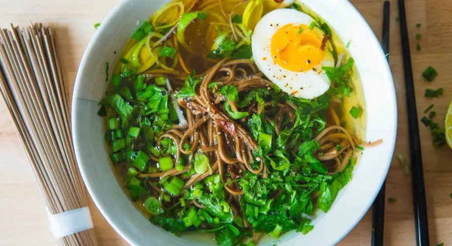A miseczce aromatyczny rosół japoński z makaronem soba, jajkiem na pół i zieloną cebulką. Obok leżą pałeczki i surowy makaron.