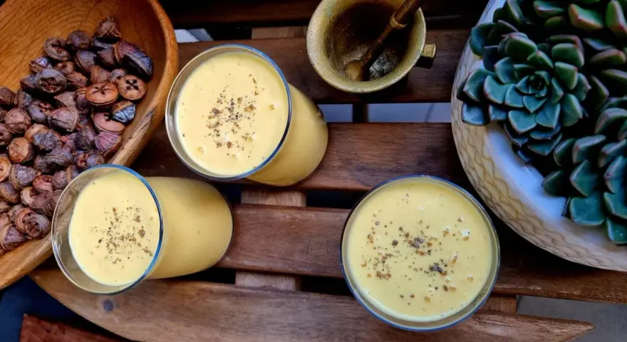Trzy szklanki kremowego mango lassi, posypane przyprawami, obok miski z suszonymi owocami i sukulentu.