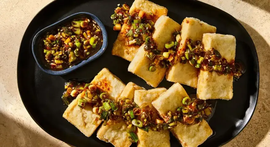 Chrupiące kawałki tofu, polane aromatycznym sosem z dymką i sezamem, podane na czarnym talerzu.