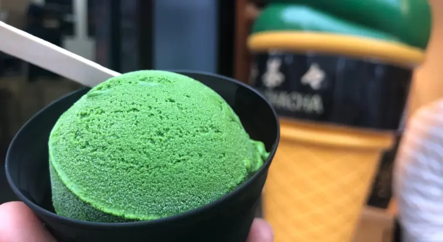 Zielone, japońskie lody matcha w czarnym kubeczku, z drewnianą łyżeczką. W tle wielki, żółty rożek z lodami.