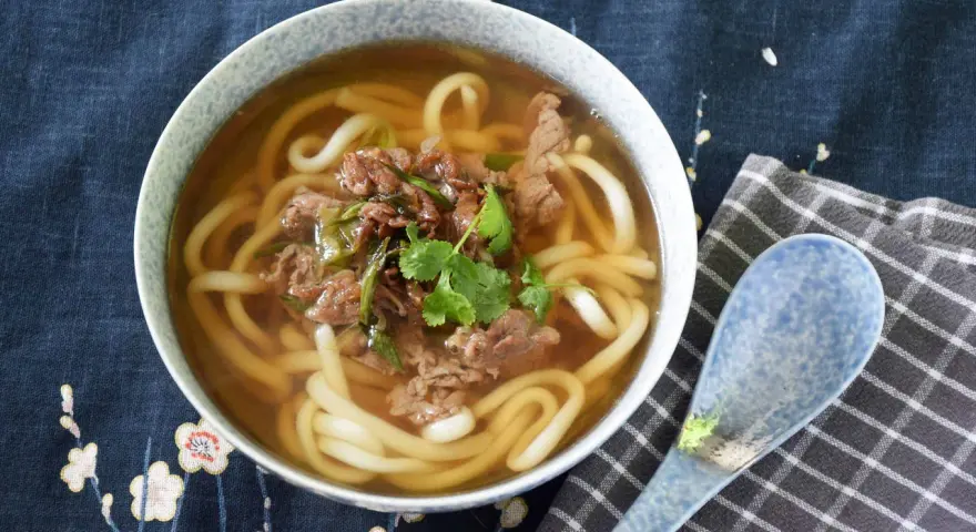 Gorąca zupa udon z wołowiną i szczypiorkiem, ozdobiona kolendrą, w misce na tle z motywem kwiatowym. Obok leży niebieska łyżka.