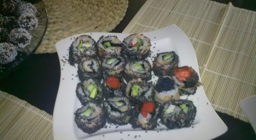 Talerz pełen sushi z sezamem, z widocznymi kawałkami awokado i ikrą. Obok deserki w wiórkach kokosowych.