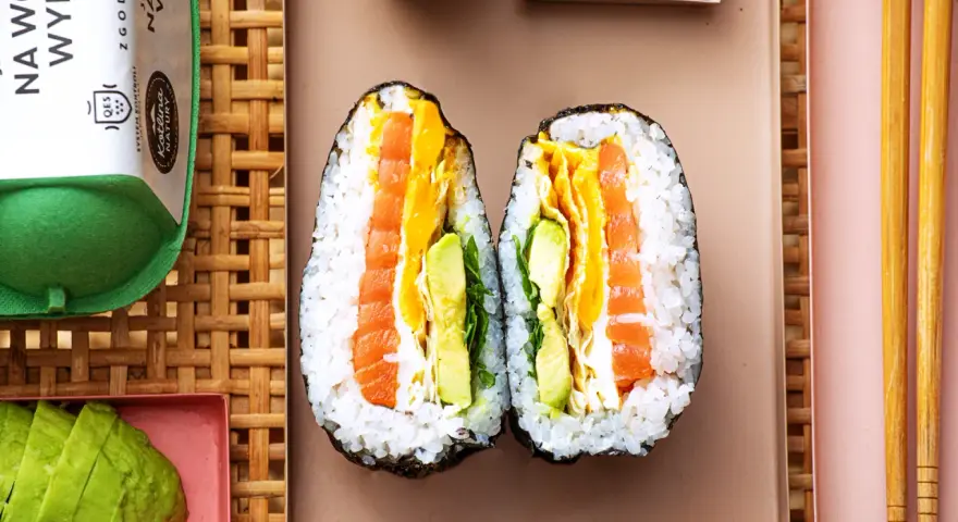 Dwa połówki onigiri z łososiem, awokado i jajkiem, podane na talerzu z pałeczkami.