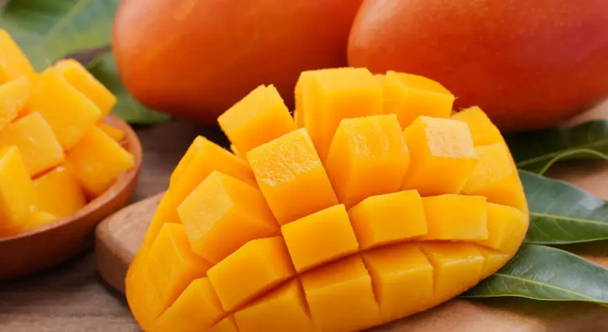 Dojrzałe mango, pokrojone w kostkę, prezentuje swoje soczyste, pomarańczowe wnętrze. Obok leżą całe owoce i liście.