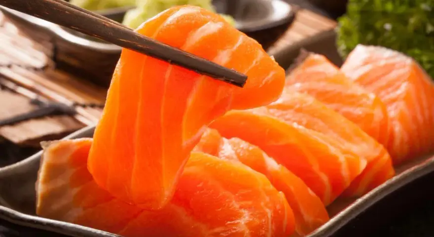 Sashimi co to? Świeże, pomarańczowe kawałki łososia podniesione pałeczkami, obok zielonej pasty wasabi i natki pietruszki.
