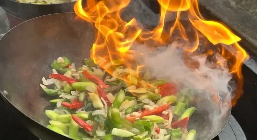 Warzywa w woku płoną w ogniu, tworząc aromatyczny stir fry. W tle widać drugie danie.
