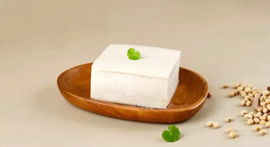 Kostka białego tofu na drewnianej miseczce, ozdobiona listkiem. Obok garść ziaren soi. To właśnie co to tofu – produkt z soi.