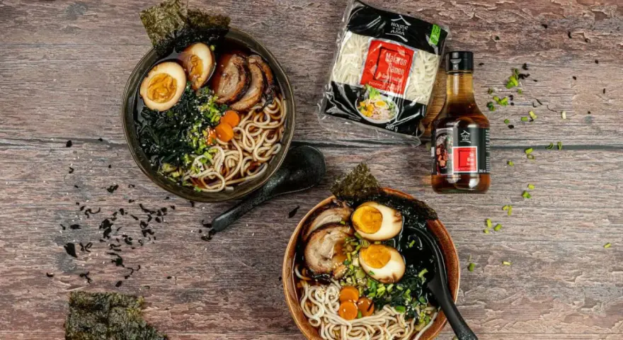 Dwa miski aromatycznego ramenu z boczkiem, jajkiem i nori. Obok opakowanie makaronu i sosu.