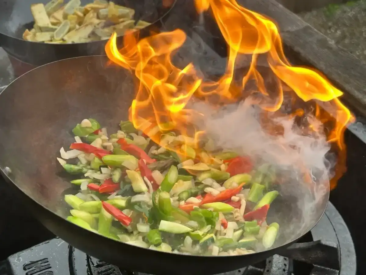 Warzywa w woku płoną w ogniu, tworząc aromatyczny stir fry. W tle widać drugie danie.