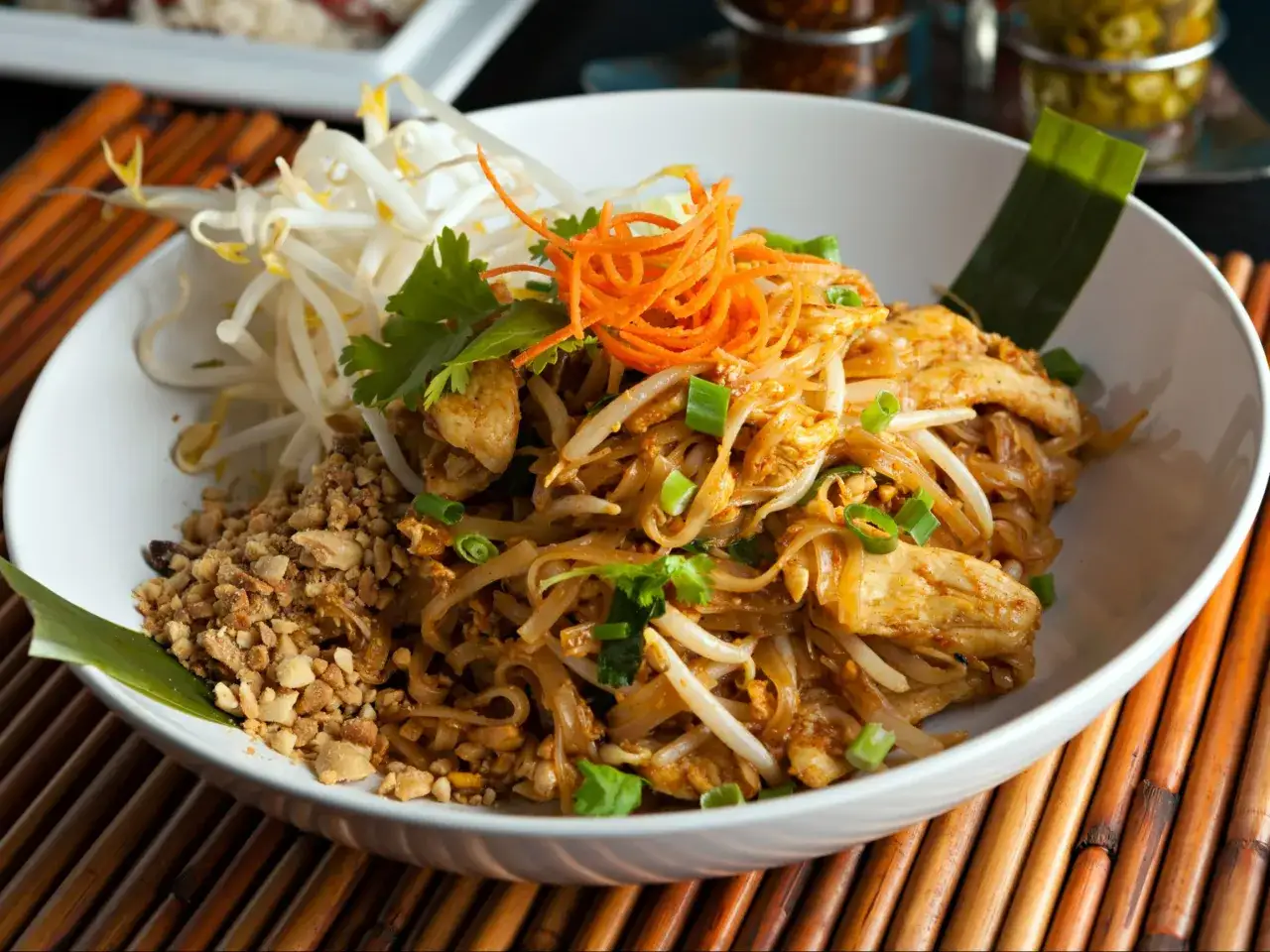 Pyszne pad thai z jajkiem, kurczakiem, kiełkami fasoli mung, orzeszkami ziemnymi i startą marchewką, podane na białym talerzu.