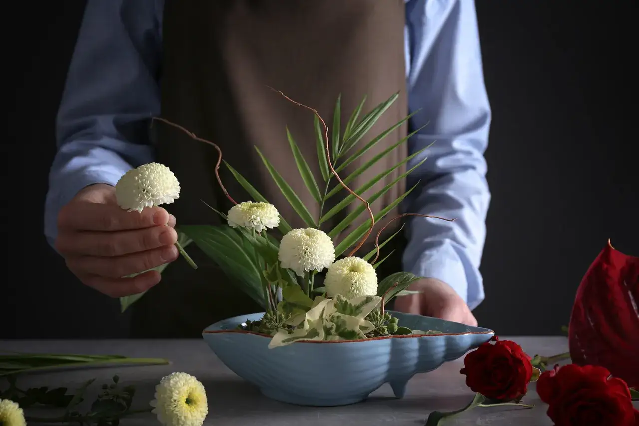 Mistrz tworzy kompozycje ikebana, dodając białe chryzantemy do błękitnej wazy.