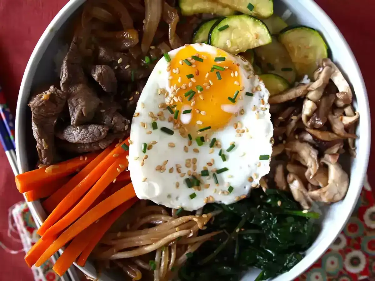 Apetyczne koreańskie jedzenie: miska bibimbap z wołowiną, warzywami, grzybami i jajkiem sadzonym, posypana sezamem.