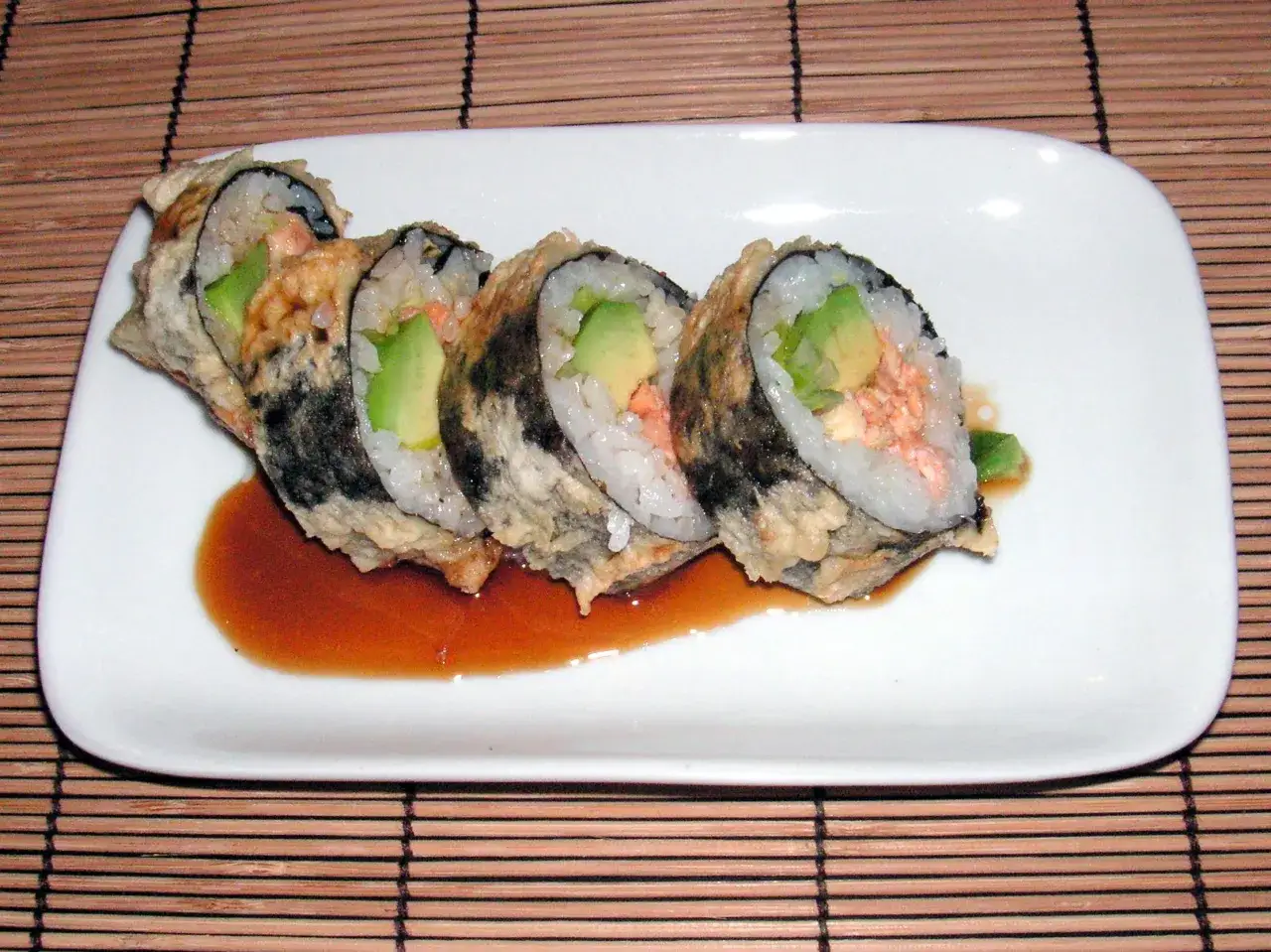 Pięć kawałków sushi z łososiem w tempurze, z awokado i ryżem, podane na białym talerzu z sosem.