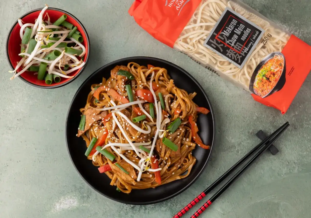Pyszny makaron z kurczakiem po azjatycku, z papryką, szczypiorkiem i kiełkami fasoli mung. Obok opakowanie makaronu chow mein.