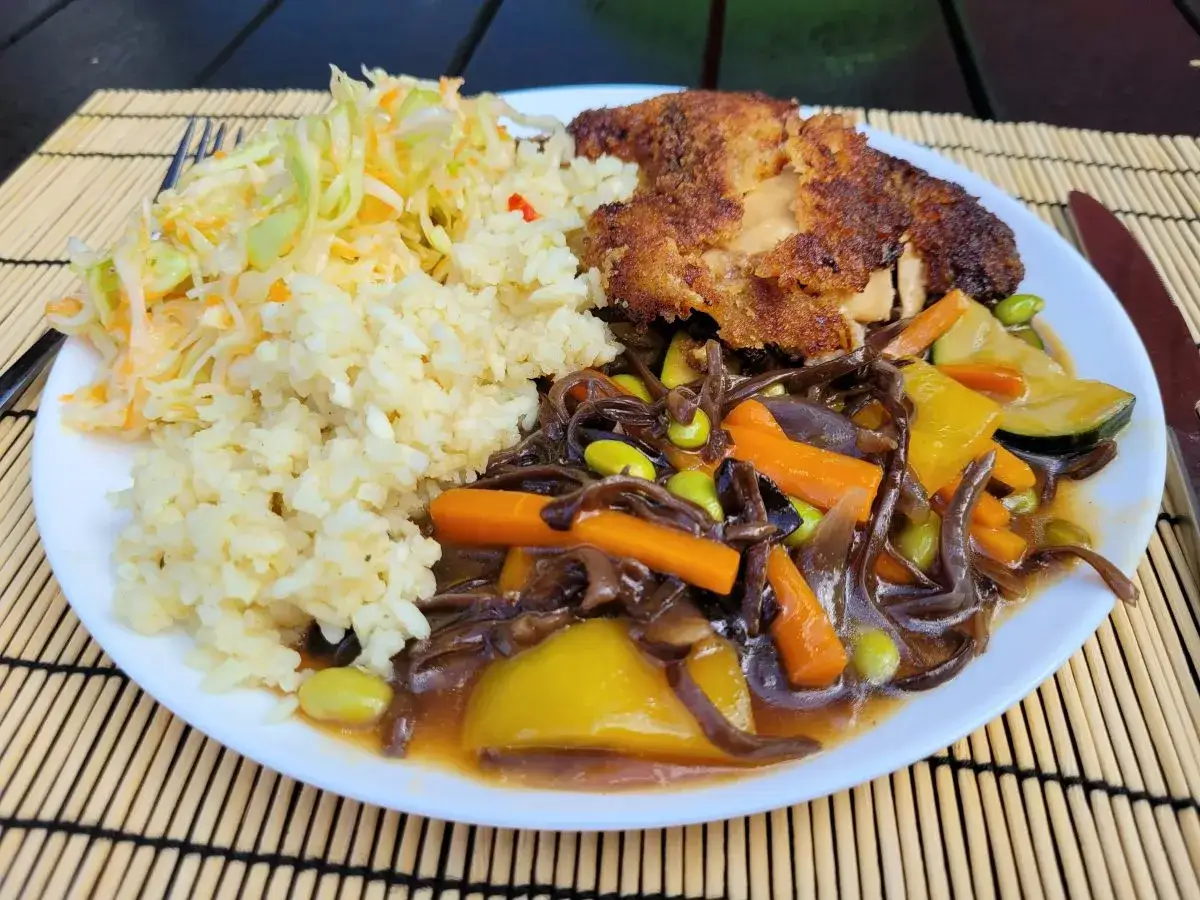Talerz z ryżem, surówką i panierowanym kotletem, obok kolorowe warzywa w sosie sojowym: marchewka, cukinia, papryka i edamame.