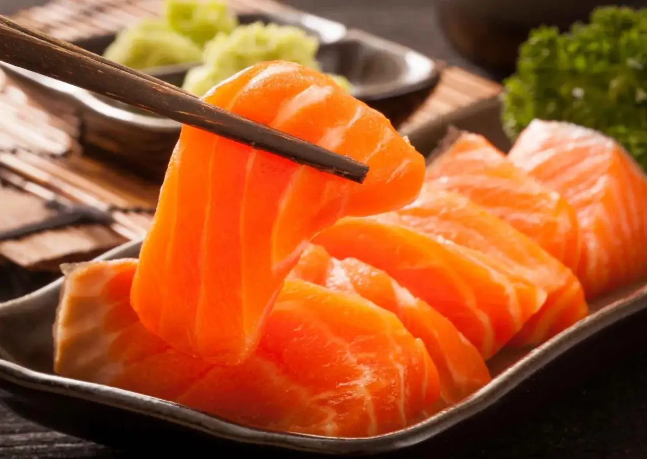 Paleczki do sushi podnoszą kawałek soczystego łososia. Obok widać talerz z plastrami sashimi, wasabi i natką pietruszki.