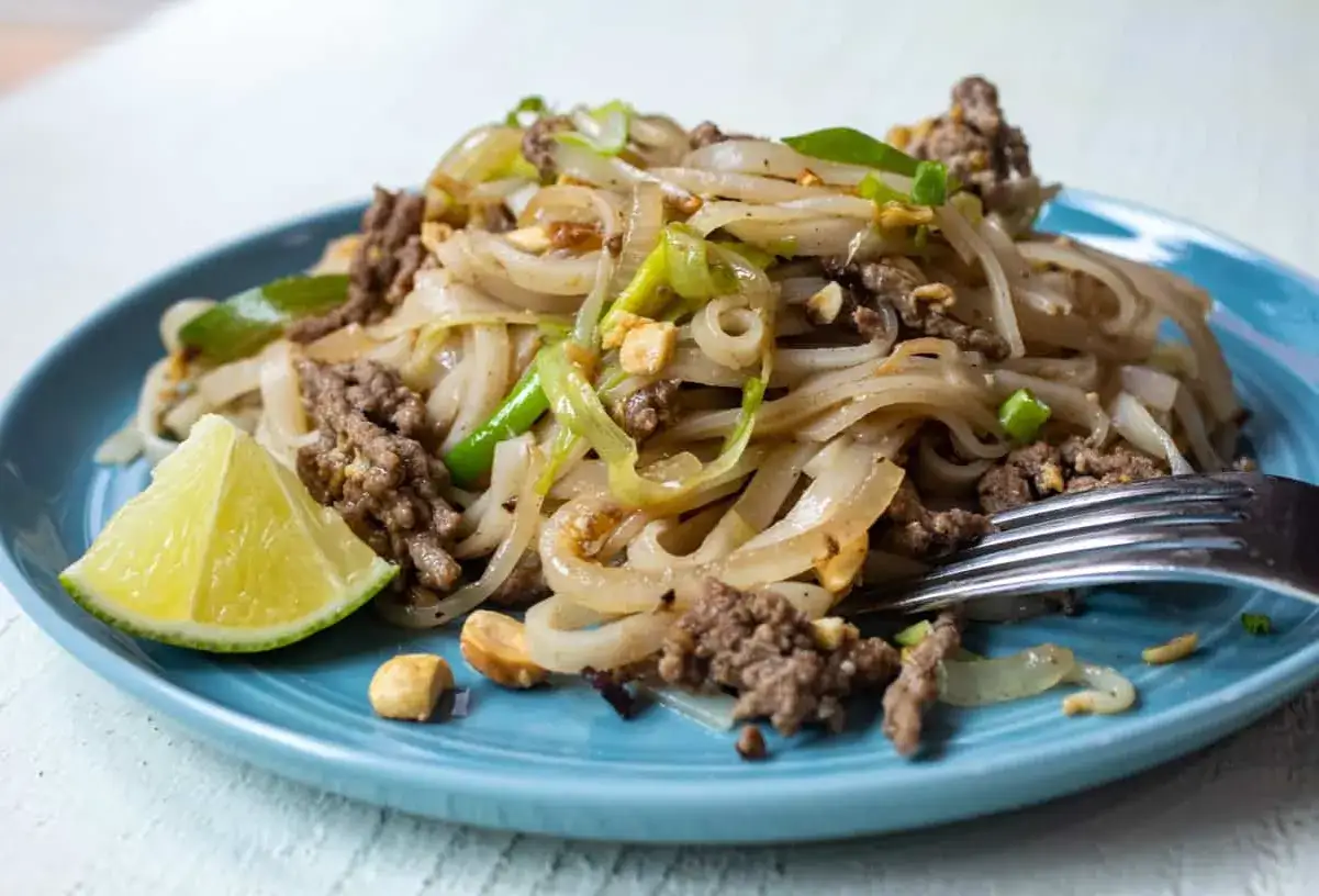 Pyszne pad thai z wołowiną, makaronem ryżowym, orzeszkami ziemnymi i ćwiartką limonki na niebieskim talerzu.