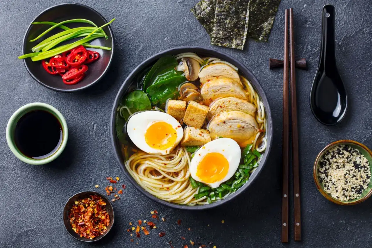 Pyszna zupa ramen z kurczakiem, jajkiem na pół, tofu i warzywami. Obok miseczka z sosem sojowym i chili.