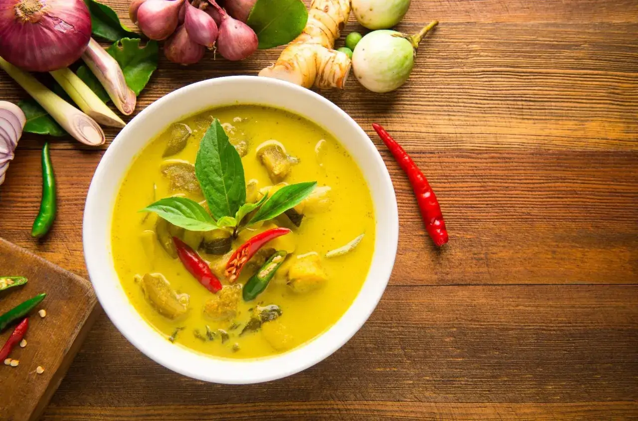 Aksamitne, żółte tajskie curry z tofu, ozdobione świeżą bazylią i papryczkami chili, otoczone składnikami: szalotką, trawą cytrynową, imbirem i bakłażanem.