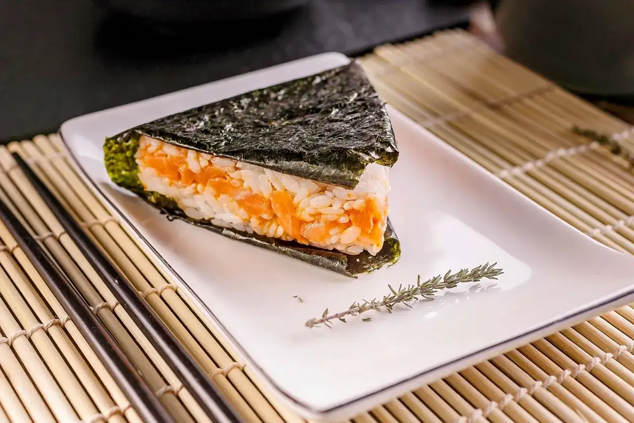 Trójkątne onigiri z łososiem, owinięte w nori, na białym talerzu z gałązką rozmarynu. Obok pałeczki i mata bambusowa.