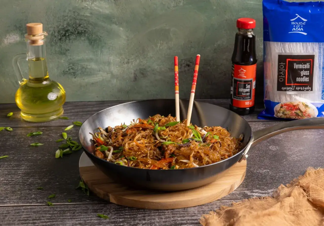 Wok z makaronem vermicelli i warzywami, obok sos ostrygowy i opakowanie makaronu. Inspiracja na makaron vermicelli przepisy.