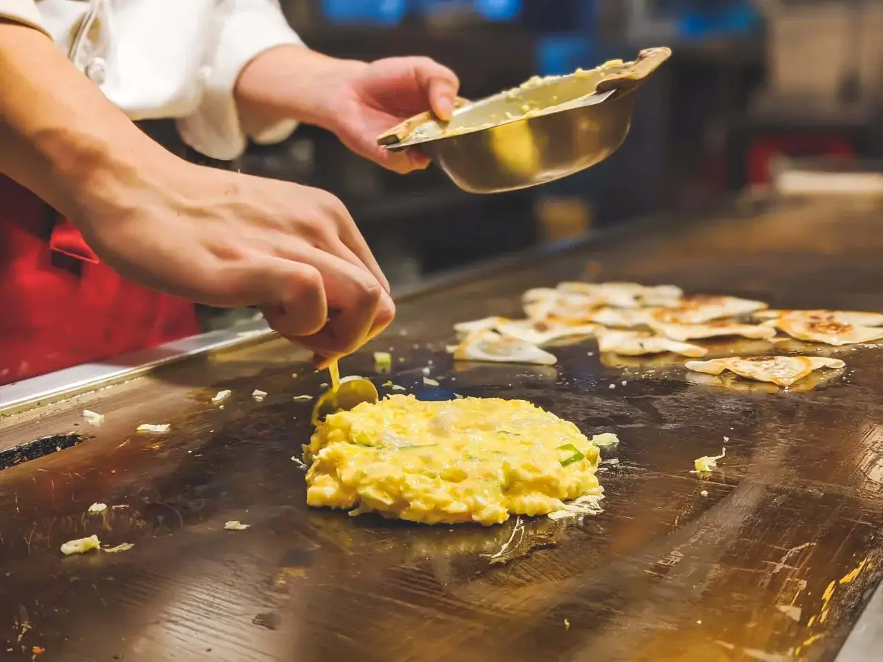 Szef kuchni formuje na gorącej płycie puszyste placki okonomiyaki, dodając składniki z metalowej miseczki.
