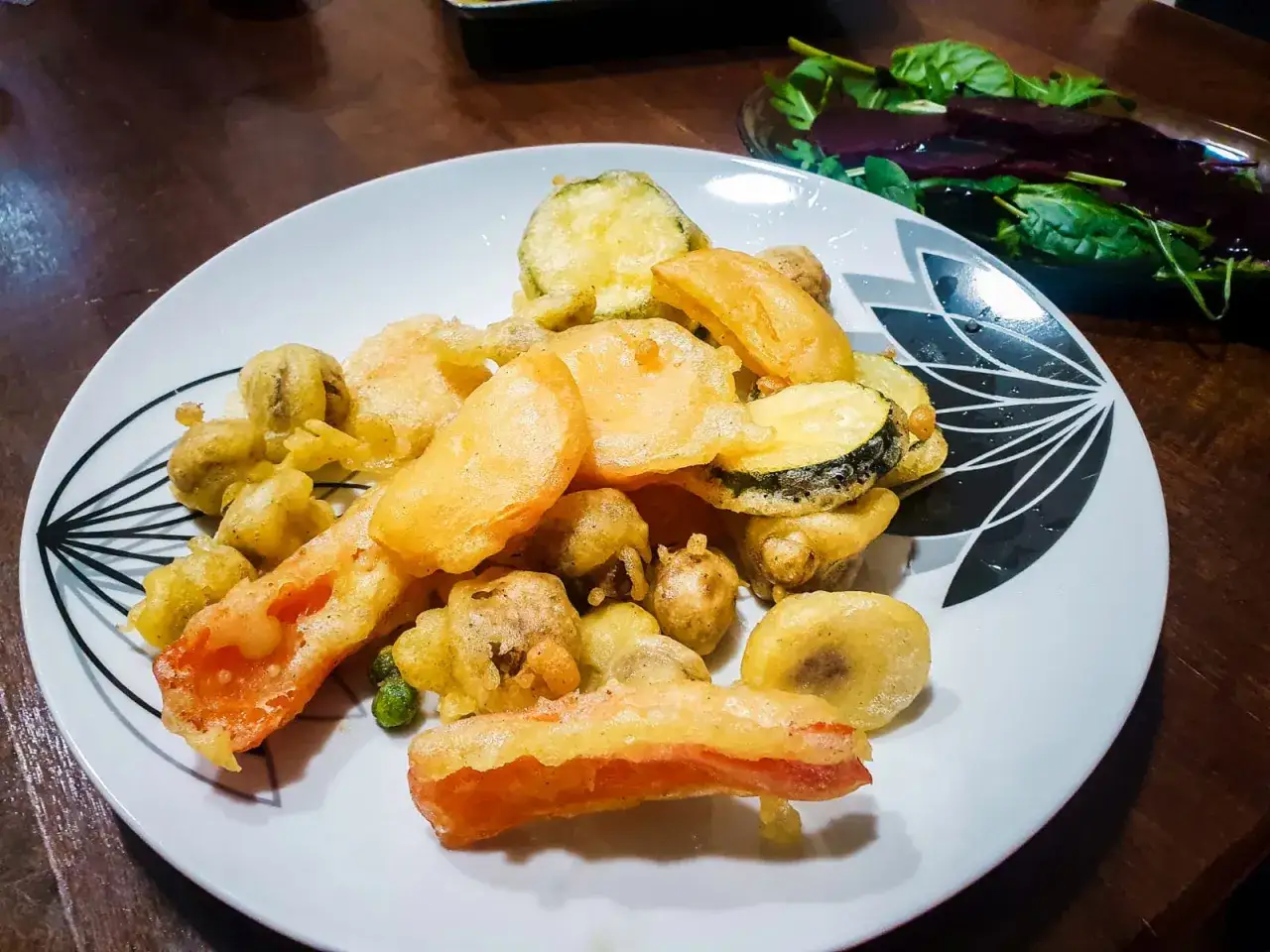 Chrupiące warzywa w tempurze: cukinia, marchewka i pieczarki, podane na białym talerzu z czarnym wzorem.