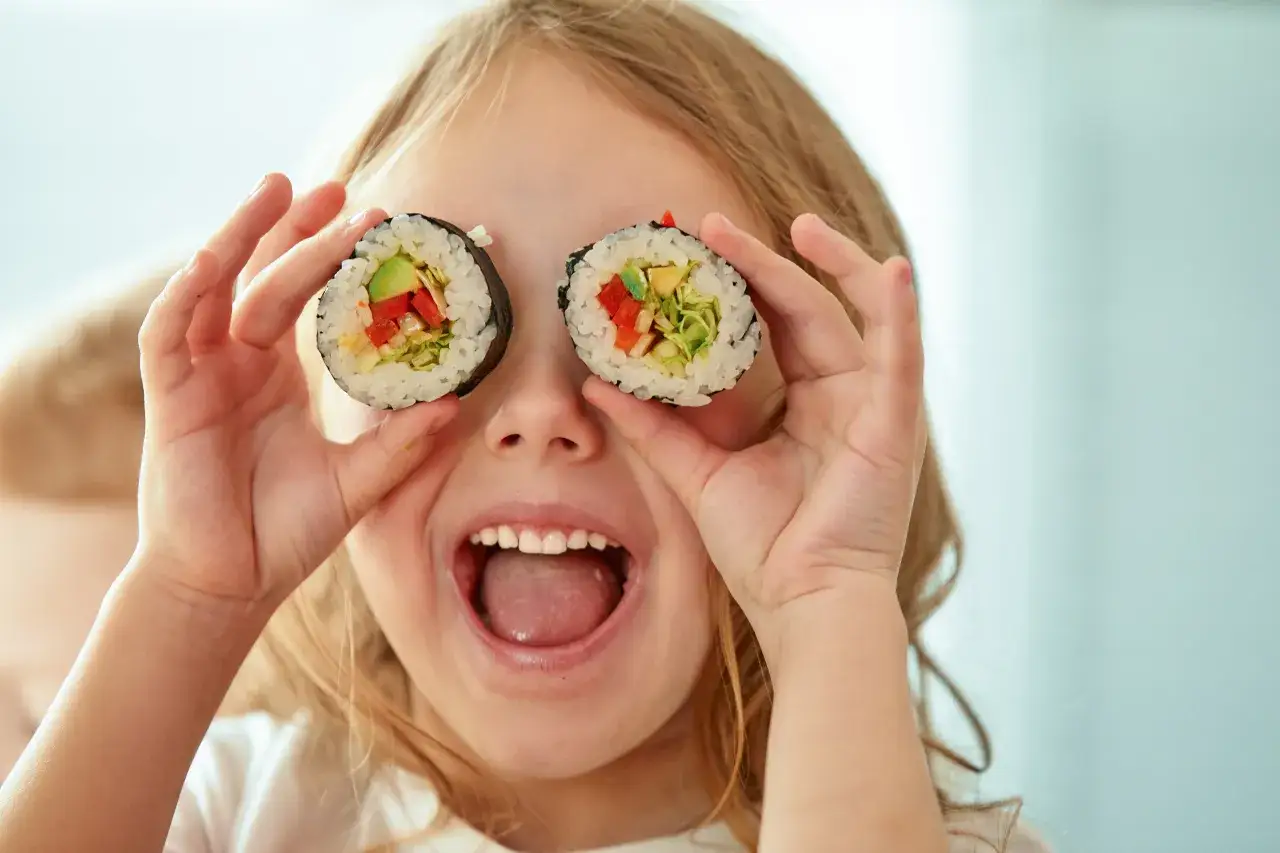 Uśmiechnięta dziewczynka trzyma kawałki sushi dla dzieci przed oczami, tworząc zabawny wyraz twarzy.