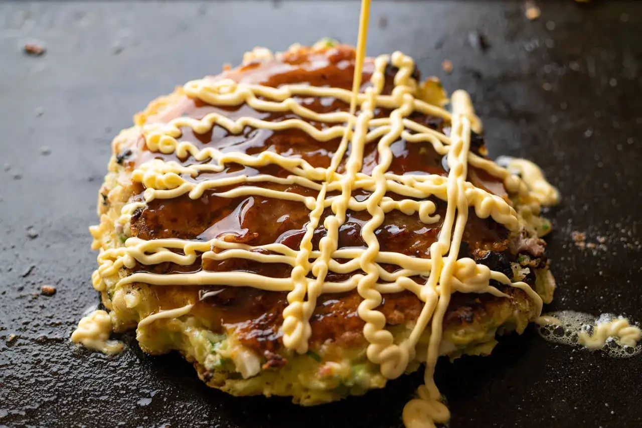 Okonomiyaki polane sosem i majonezem japońskim, przygotowywane na gorącej płycie.