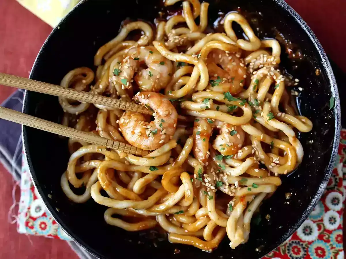 Pyszne udon z krewetkami w sosie, posypane sezamem i natką pietruszki. Danie prezentowane w czarnej misce, podane z pałeczkami.