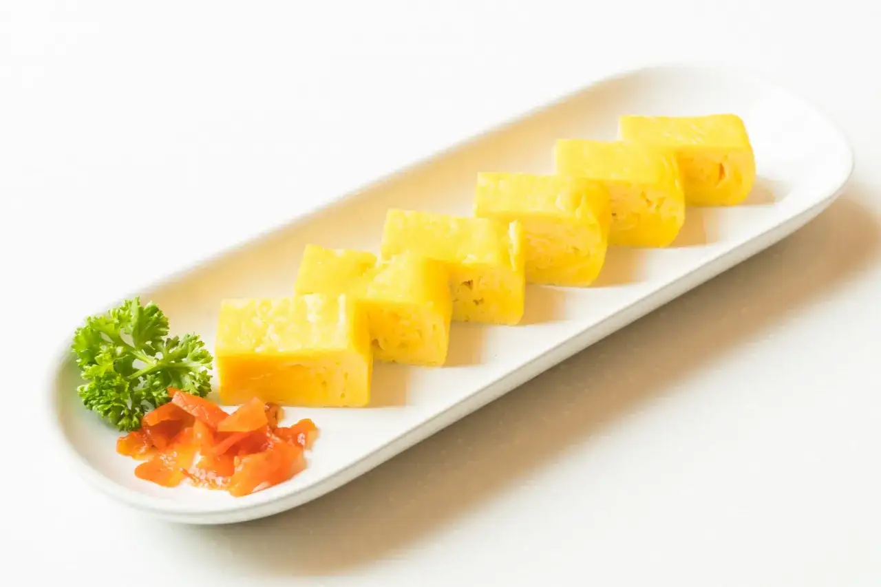 Sześć kawałków żółtego, puszystego sushi w omlecie, podanych z natką pietruszki i pomarańczowym imbirem na białej tacy.