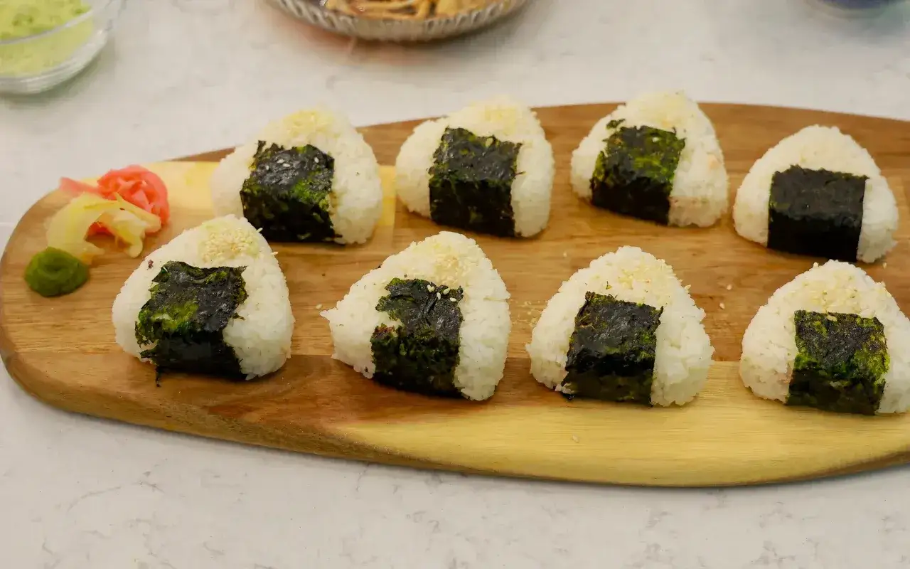 Na desce leży osiem trójkątnych onigiri z tuńczykiem, owiniętych w nori, posypanych sezamem. Obok znajduje się wasabi i marynowany imbir.