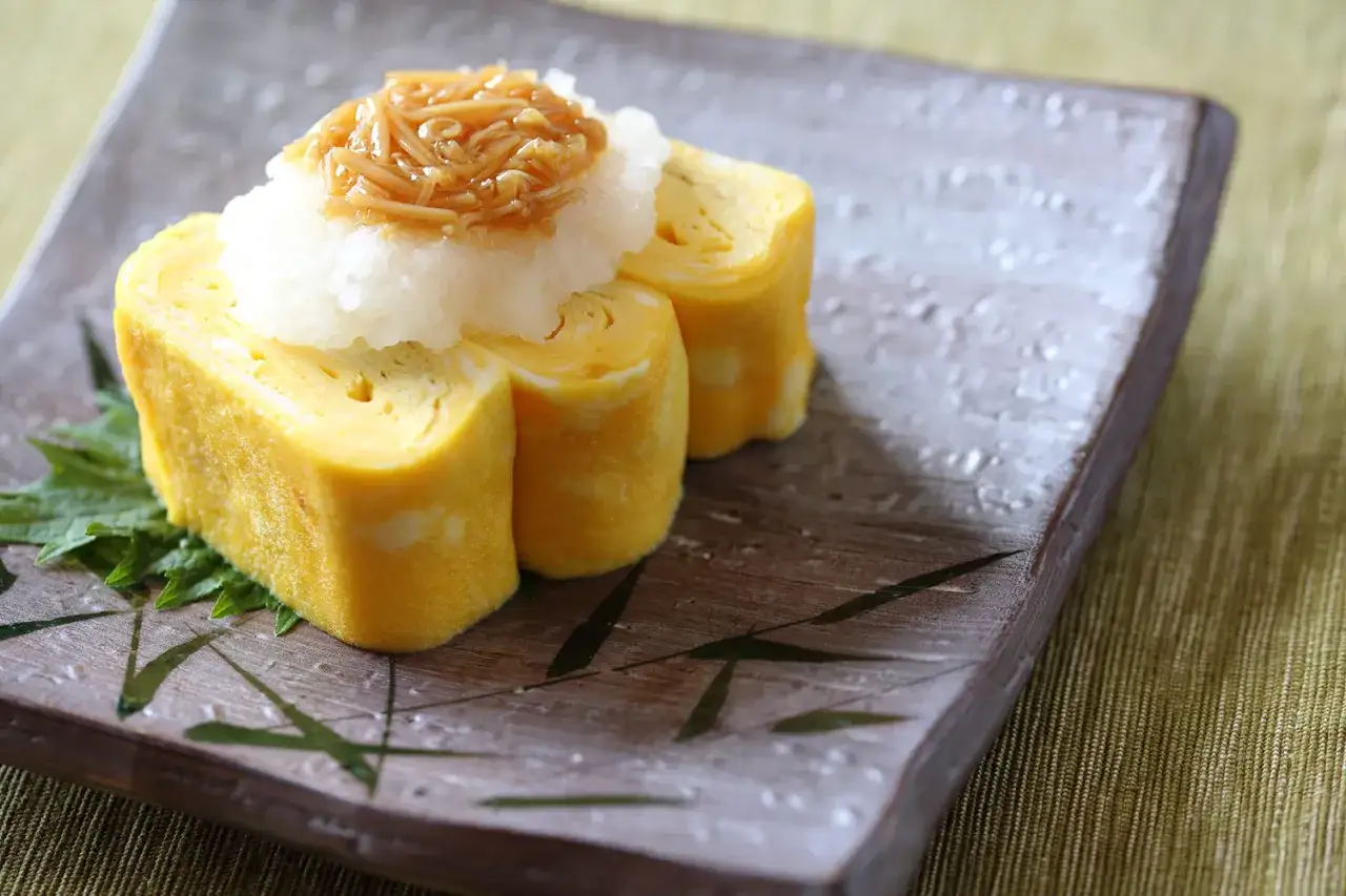 Trzy kawałki tamagoyaki, czyli japońskiego omletu, ułożone jak nigiri. Na wierzchu starta rzodkiew i grzyby enoki.