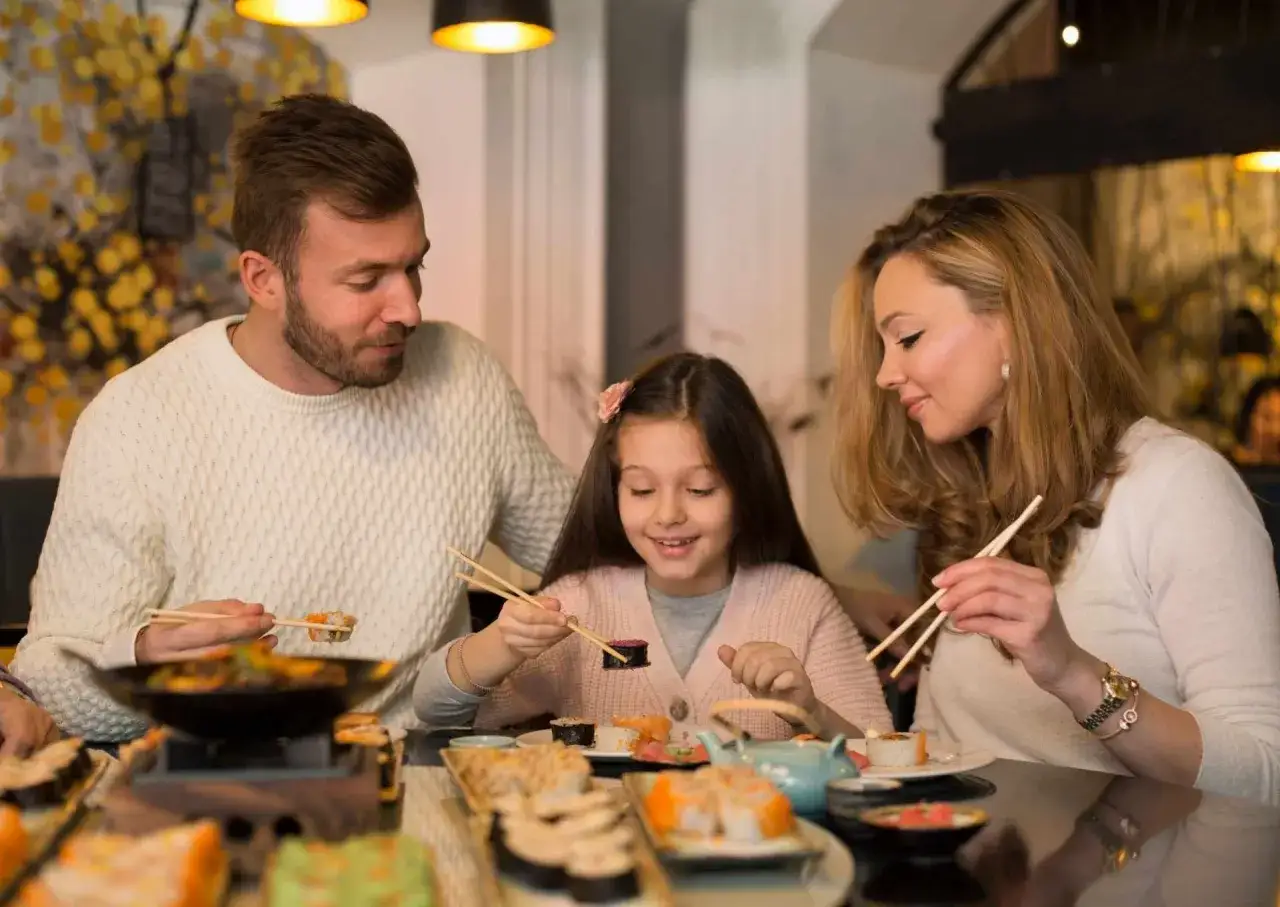 Rodzina delektuje się pysznym sushi. Dziewczynka z uśmiechem próbuje swoich pierwszych roladek, a rodzice z dumą obserwują, jak smakuje im sushi dla dzieci.