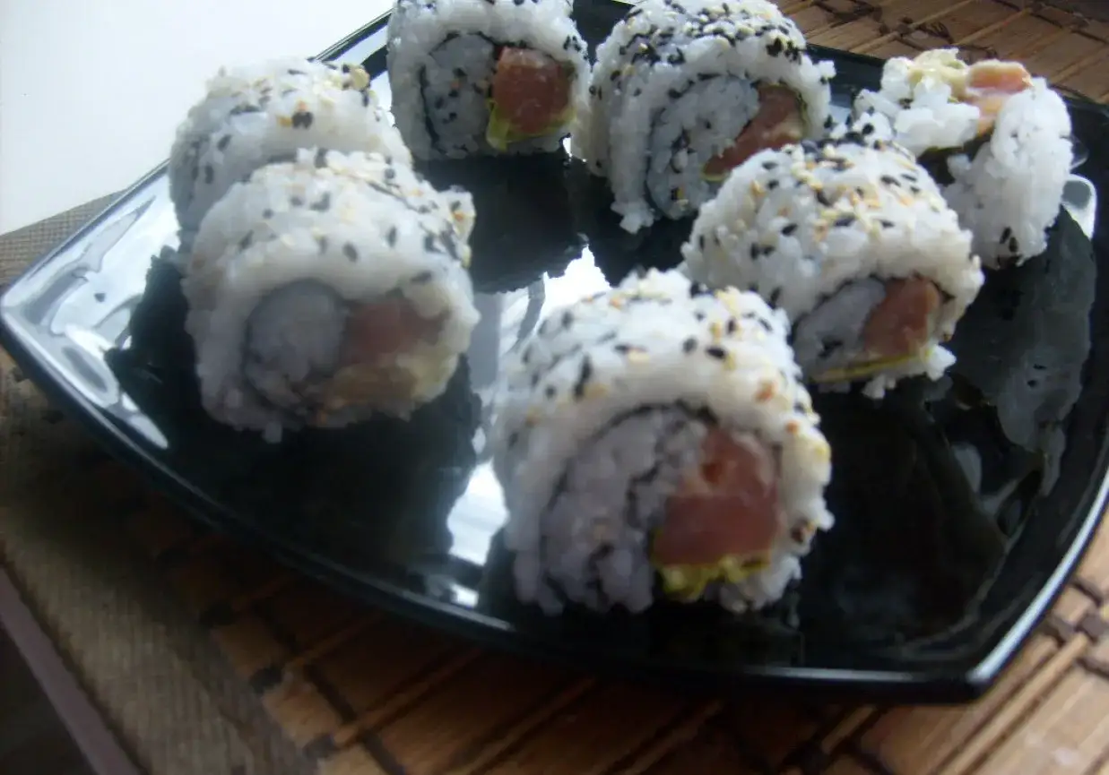 Kilka kawałków sushi uramaki z łososiem i awokado, posypane sezamem, na czarnym talerzu.